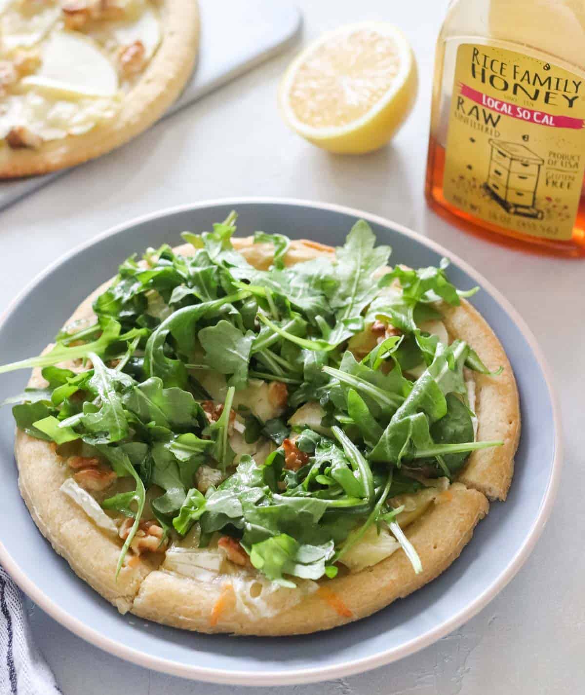 personal pizza topped with arugula on a blue plate next to a bottle of honey, a cut lemon, and a cutting board with another apple brie pizza