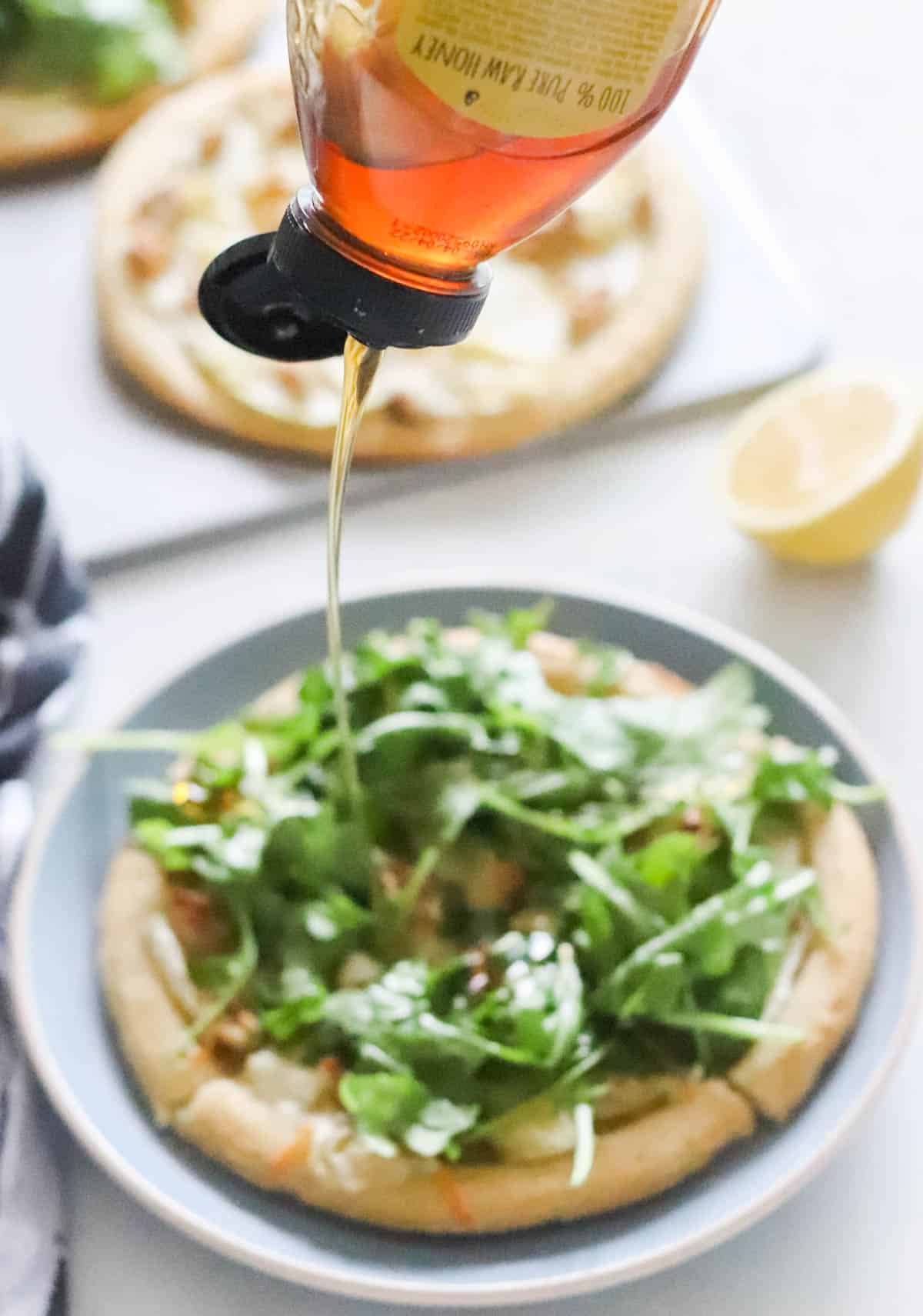 honey being squeezed onto a personal pizza topped with arugula on a blue plate with a pizza and a cut lemon in background