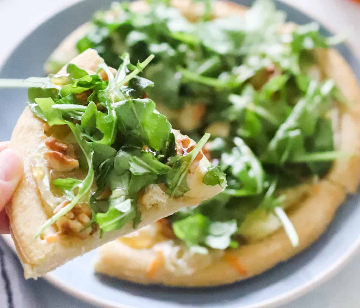 hand holding a piece of pizza topped with walnuts and arugula with a plate of pizza in background