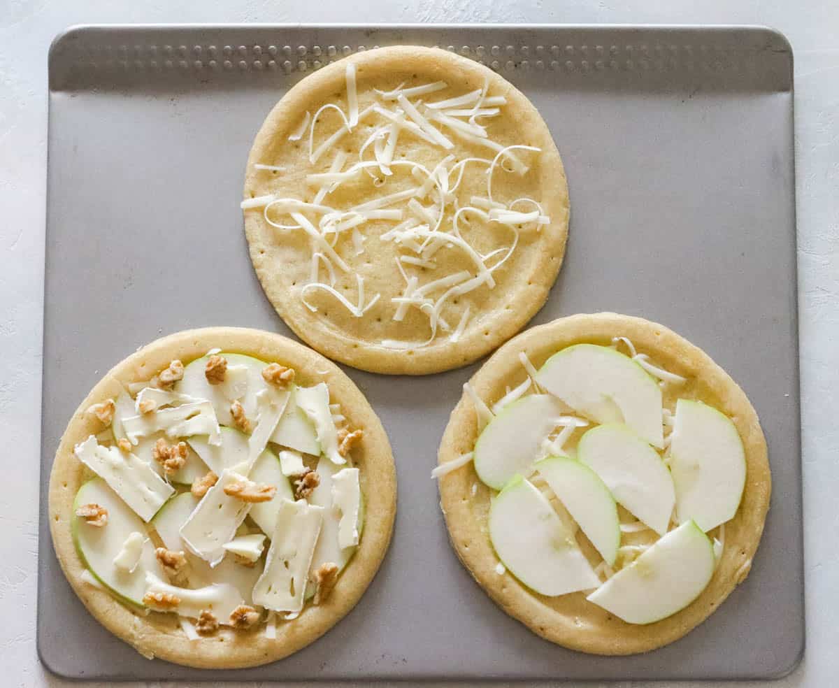 silver baking sheet with three small pizza crusts, one topped with cheese, one topped with cheese and apple slices, one topped with cheese, apple slices, and walnuts
