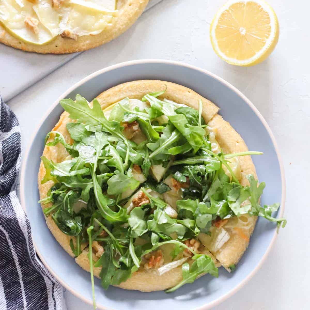 blue plate with pizza topped with arugula next to a grey napkin, half a lemon, and another apple brie pizza on a cutting board