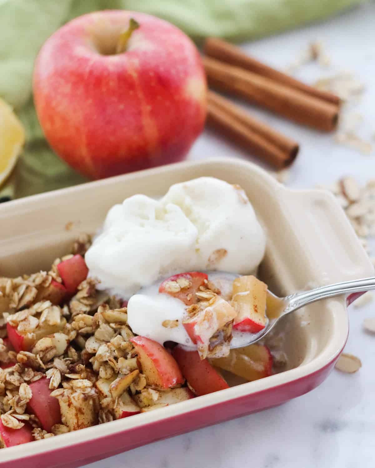 silver spoon scooping out baked apples with ice cream from a small red baking dish with apple and cinnamon sticks in background