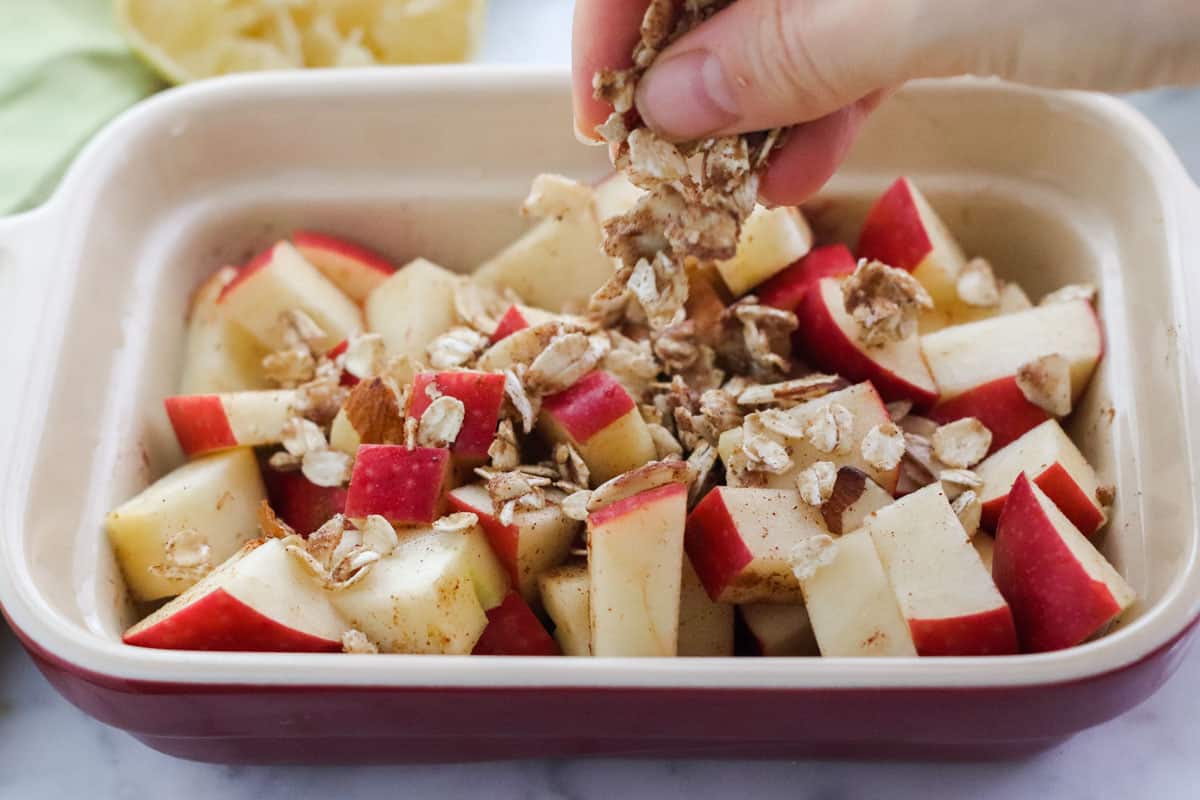 hand sprinkling oats over apples and cinnamon in a small red baking dish