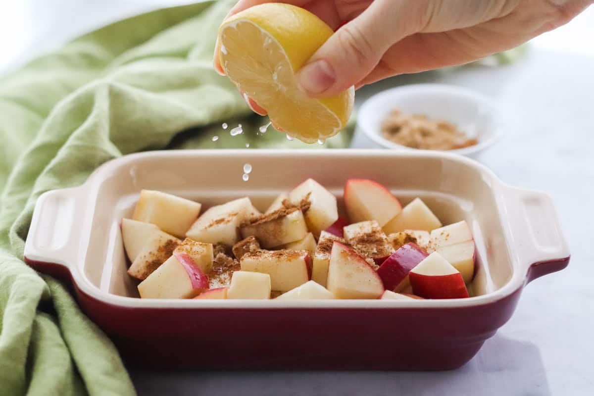 hand squeezing lemon juice over cinnamon-covered apples in a small baking dish