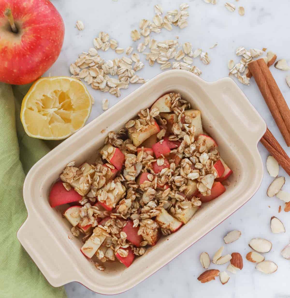 microwaved apple in small casserole dish on counter with green napkin, scattered almond slices, cinnamon stick, apple, cut lemon