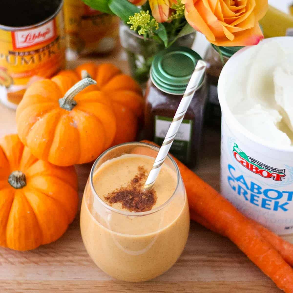 white and gold paper straw in a glass of pumpkin smoothie sprinkled with cinnamon, next two small pumpkins, carrots, container of yogurt, spices, and flowers