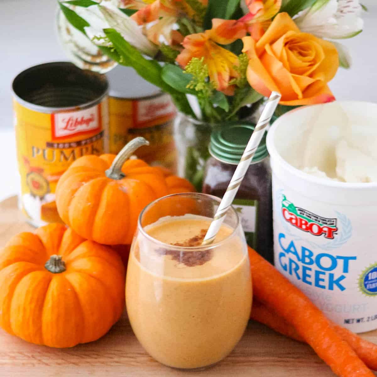 wooden counter with pumpkin pumpkin smoothie in glass with straw, next to yogurt, pumpkins, canned pumpkin puree, and orange flowers