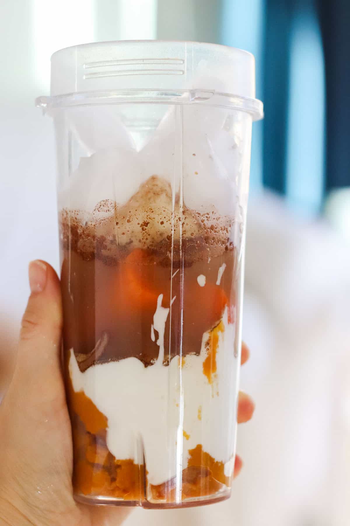 hand holding blender cup with pumpkin puree, carrots, yogurt, banana, spices, water, and ice