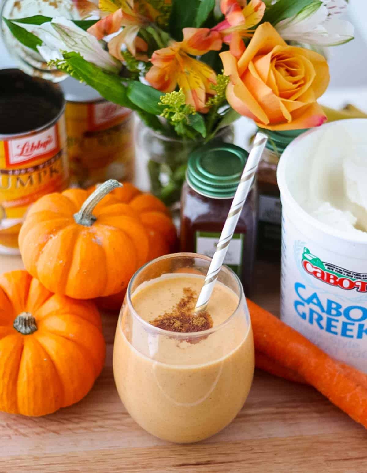 wine glass filled with pumpkin smoothie and a paper straw surrounded by mini pumpkins, can of pumpkin, flowers, spices