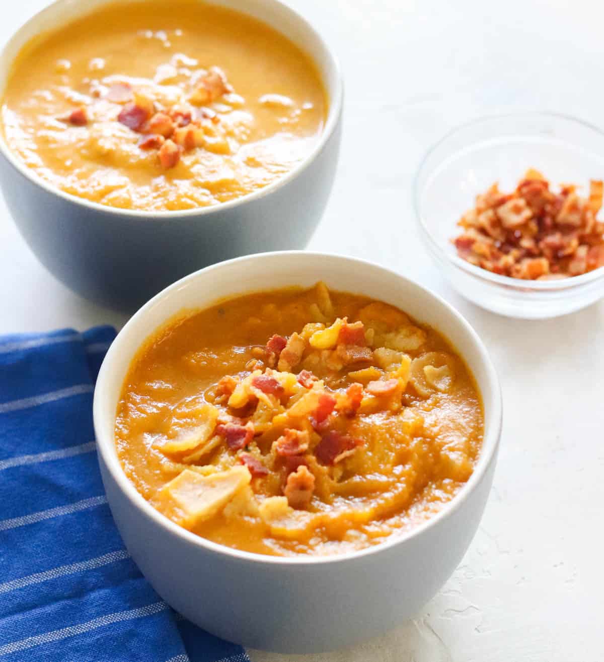 small clear bowl with bacon crumbles, two grey bowls of pumpkin soup topped with bacon, blue and white striped napkin
