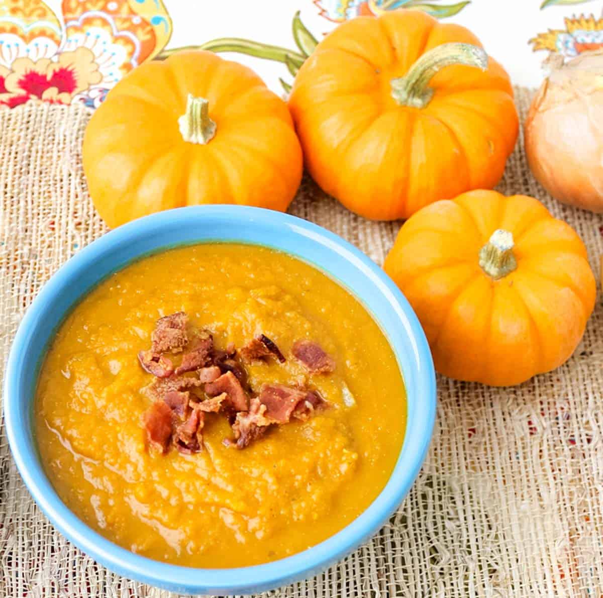 small decorative pumpkins on a patterned placemat with a blue bowl of soup topped with bacon