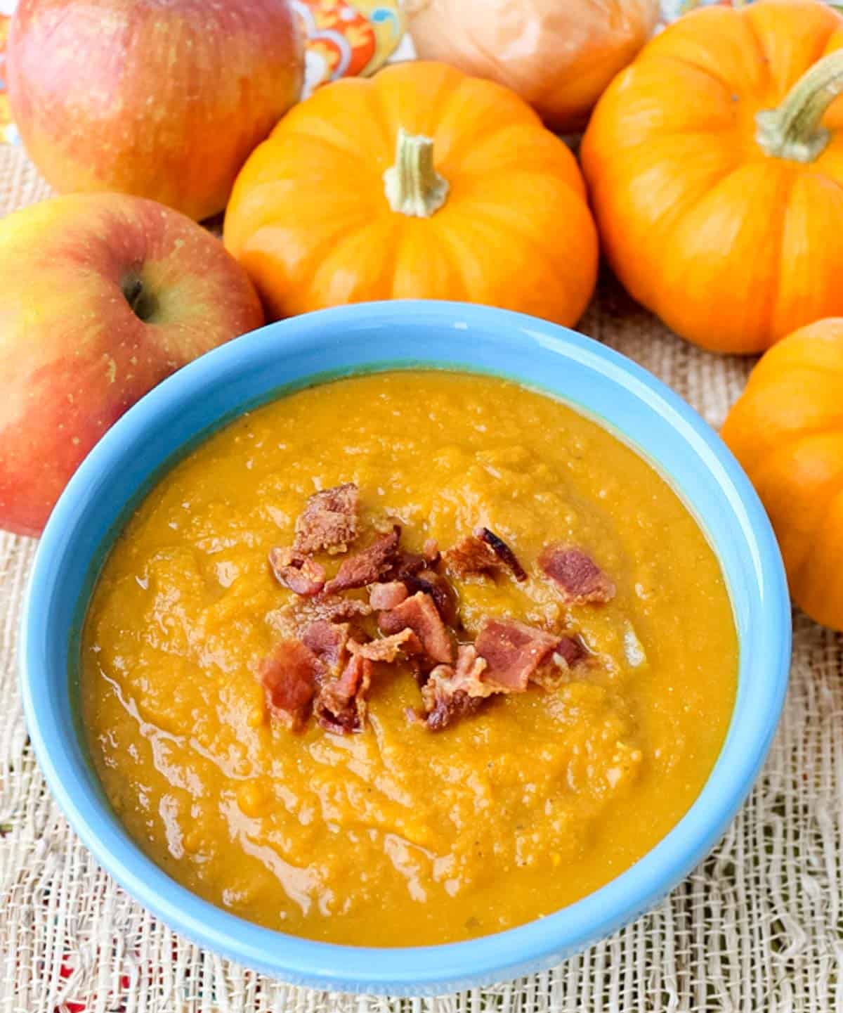 blue bowl of soup sprinkled with bacon next to apples and small pumpkins