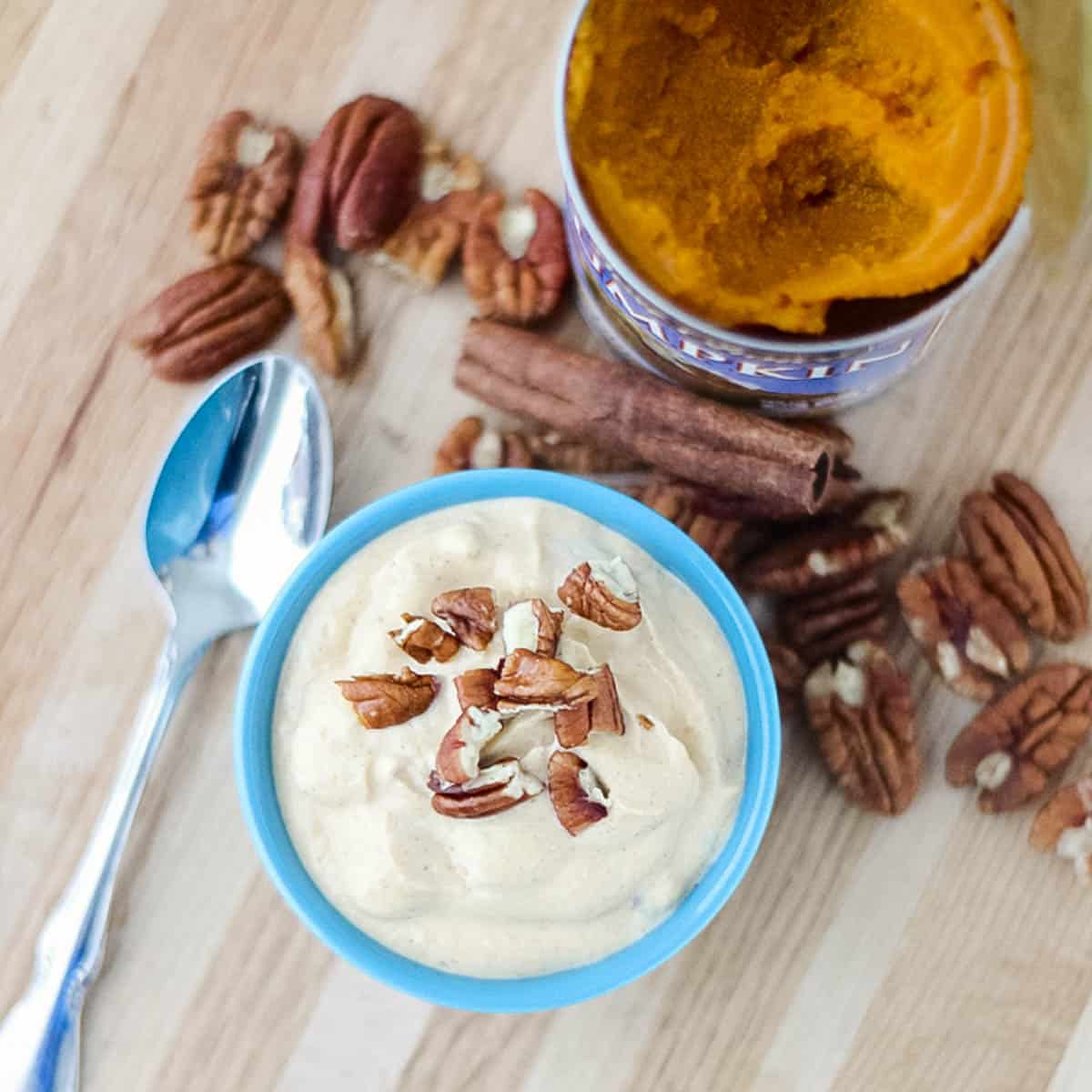 silver spoon, blue bowl of pumpkin yogurt topped with pecans on a wooden counter surrounded by pecans, cinnamon stick, and open can of pumpkin puree