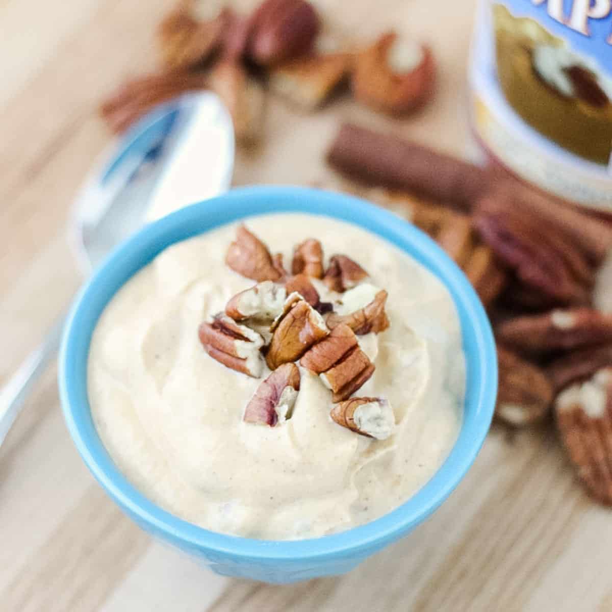 blue bowl of pumpkin yogurt topped with pecans on a wooden counter surrounded by pecans and cinnamon stick