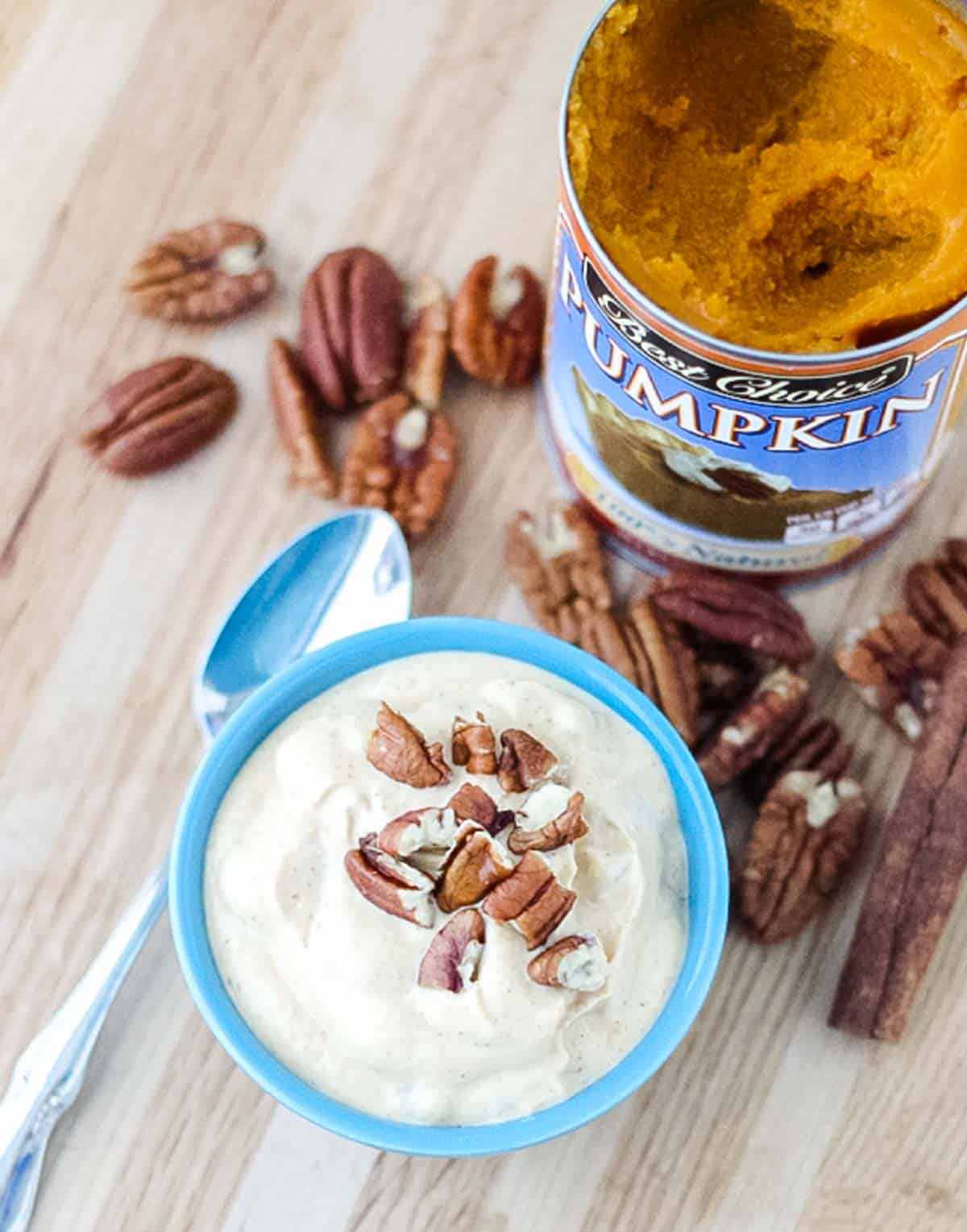 silver spoon and blue bowl of pumpkin yogurt topped with pecans on a wooden counter surrounded by pecans and open can of pumpkin puree