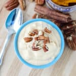blue bowl of pumpkin yogurt topped with pecans on a wooden counter with silver spoon surrounded by pecans and cinnamon stick