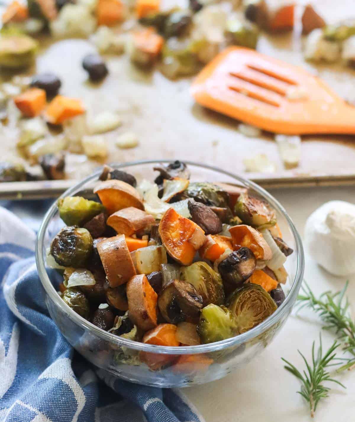 clear bowl of roasted veggies next to sprigs of rosemary, a baking sheet with more veggies, and a blue napkin