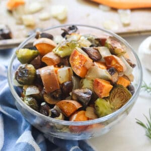 blue napkin with clear bowl with roasted sweet potatoes, brussels sprouts, onions, and mushrooms plus a baking sheet in the background