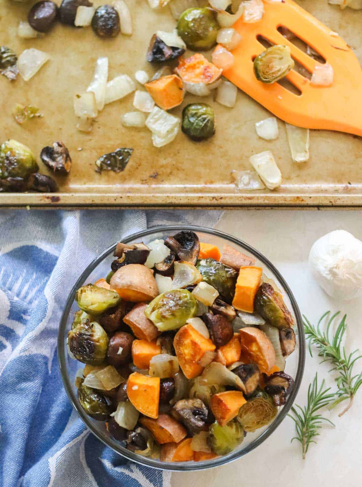 overhead view of a bowl of roasted vegetables on a blue napkin next to sprigs of rosemary and a baking sheet with extra roasted veggies