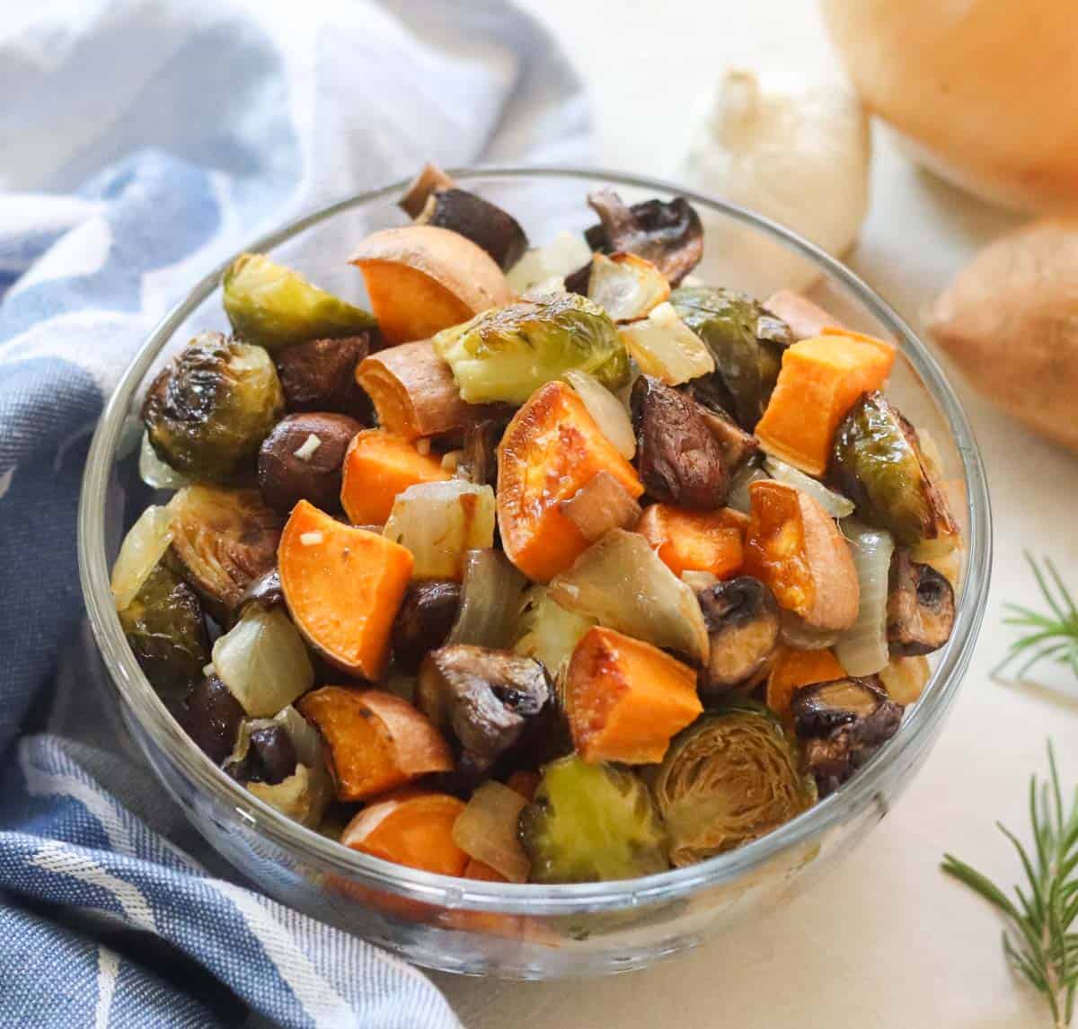 clear bowl with roasted sweet potatoes, mushrooms, onions, and brussels sprouts with rosemary, vegetables, and a blue napkin near the bowl
