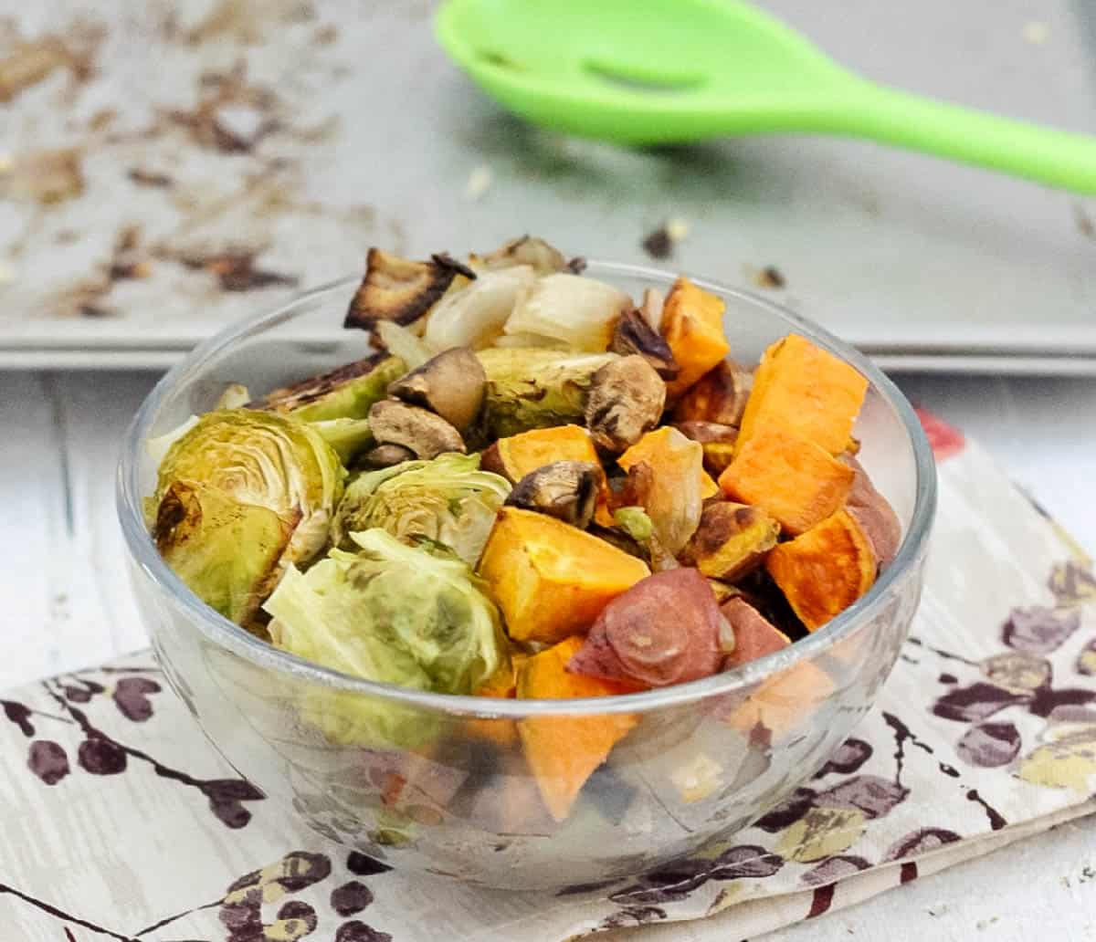 clear bowl of roasted vegetables on a floral napkin in front of an empty baking sheet