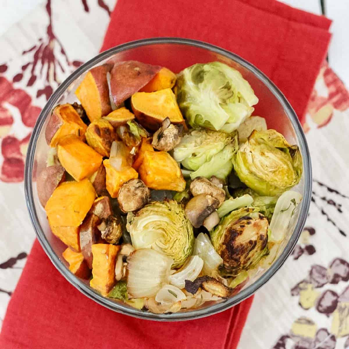 bowl of roasted onions, brussels sprouts, sweet potatoes, and mushrooms on a red napkin and a floral napkin