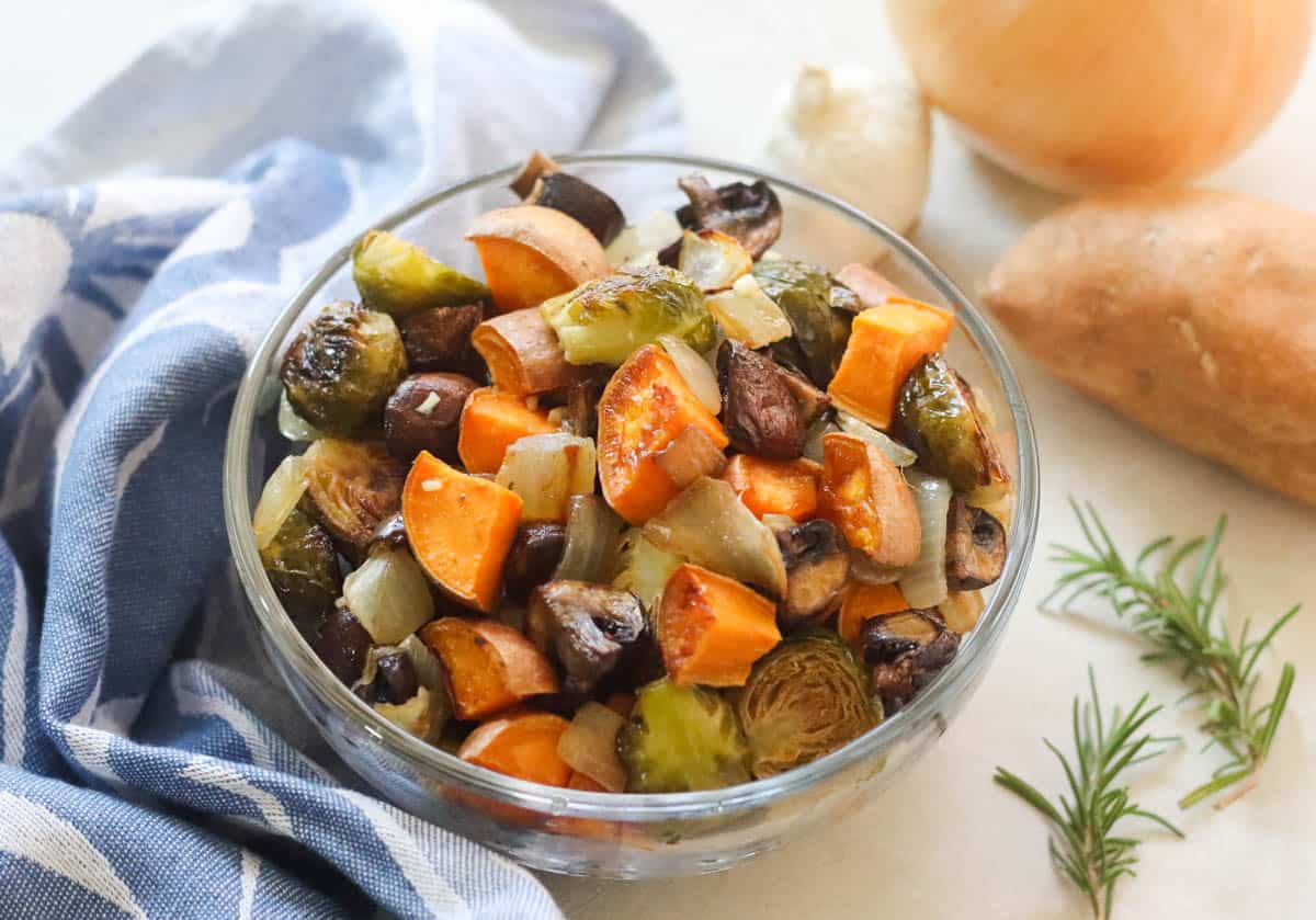 clear bowl of roasted veggies next to rosemary sprigs, sweet potato, onion, garlic, and blue napkin