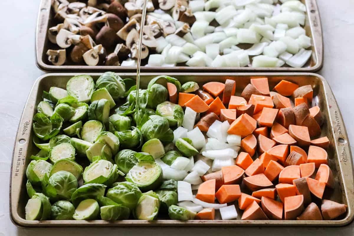 oil being poured onto brussels sprouts, onions, and sweet potatoes on a baking sheet