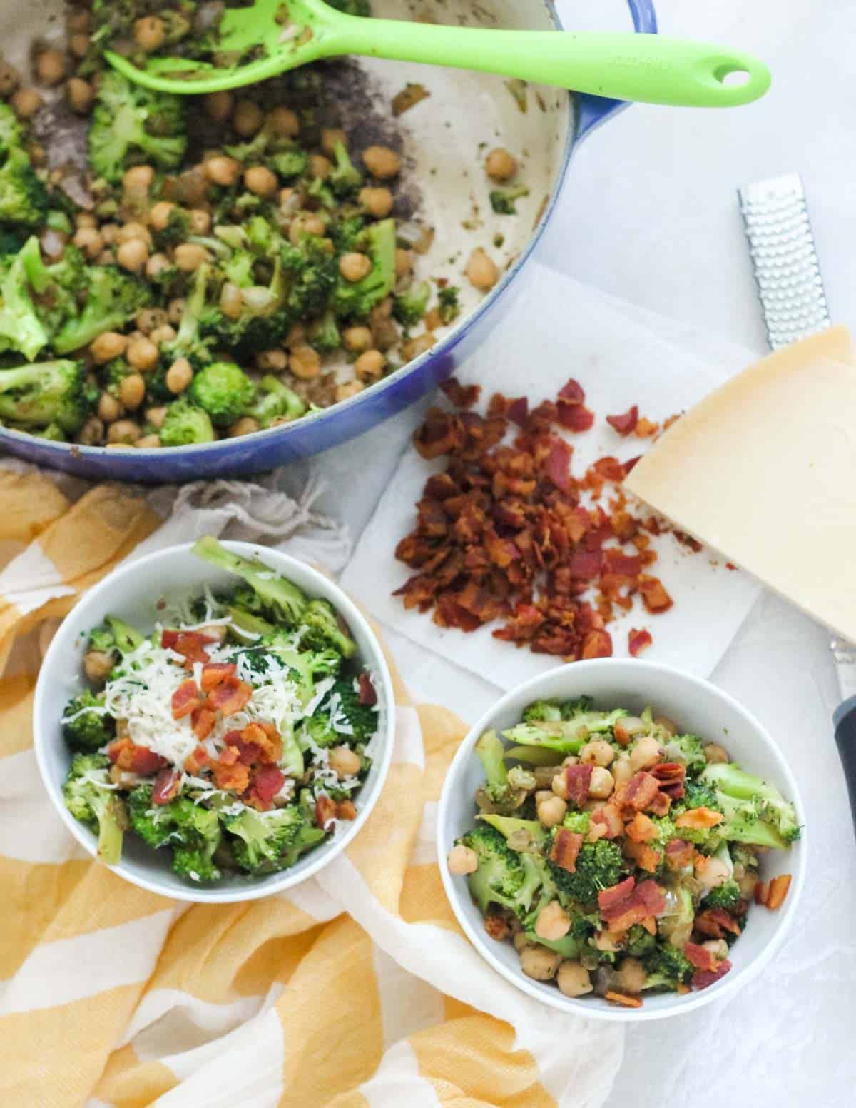 two bowls with chickpeas, broccoli, and bacon next to a pile of bacon, wedge of parmesan, and a blue pot with broccoli