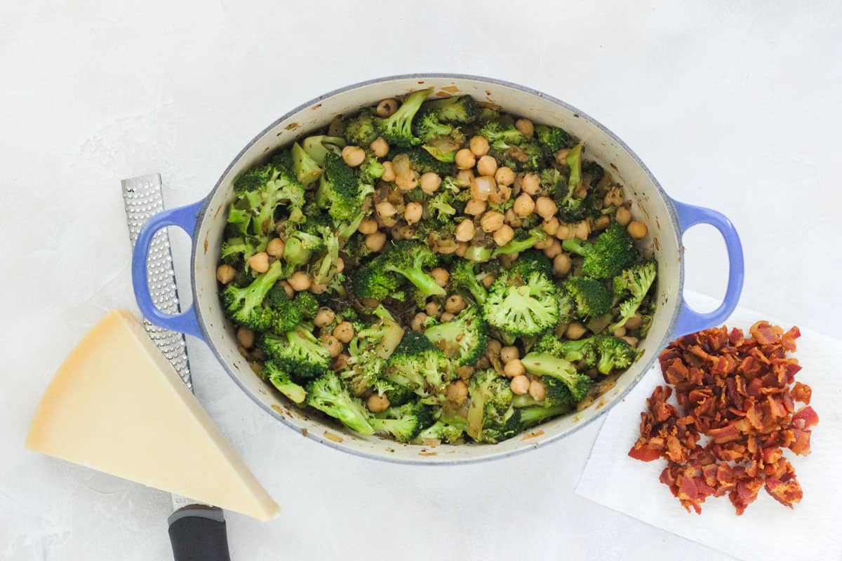 blue pot with broccoli and chickpeas next to a microwave with a wedge of parmesan and a napkin with cooked bacon