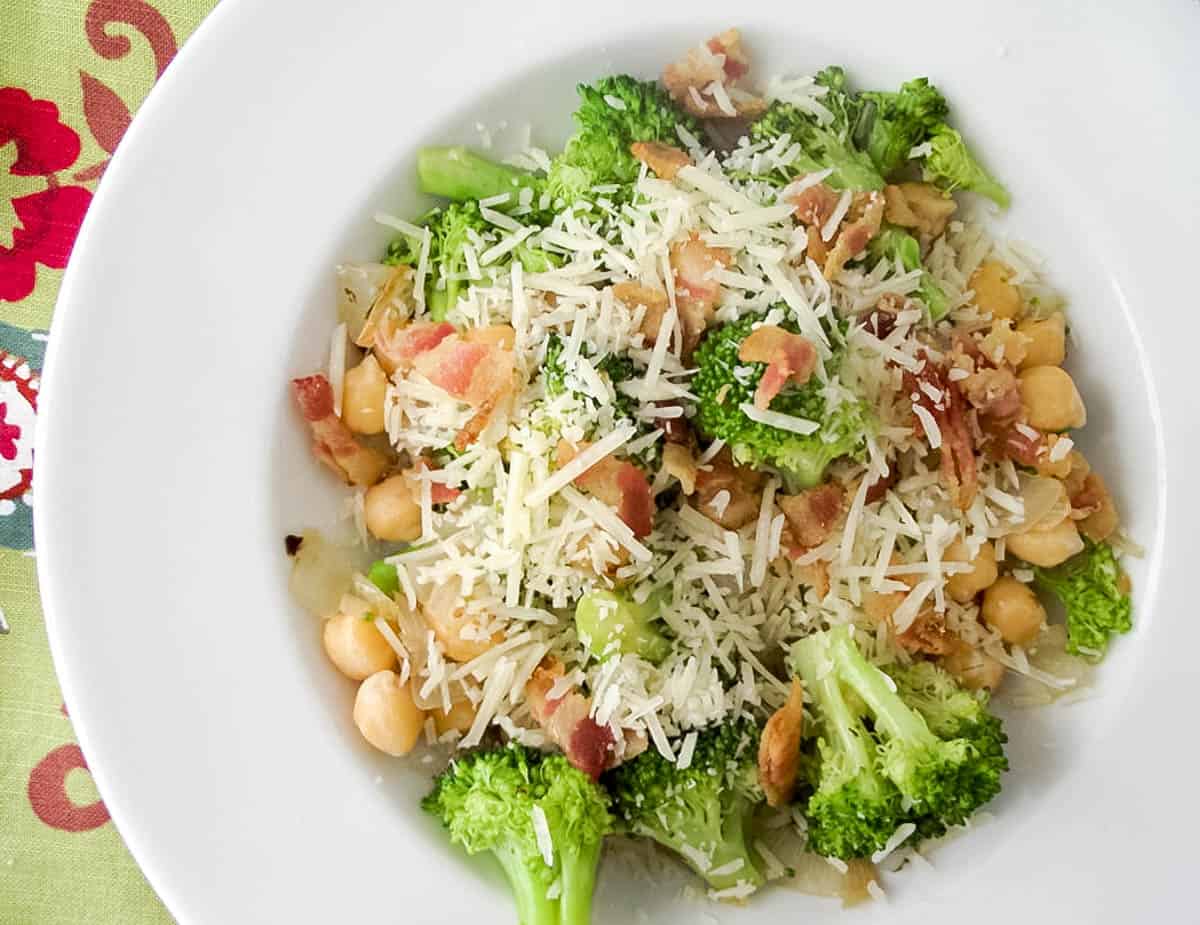 broccoli tossed with chickpeas and onions topped with bacon and shredded parmesan in a white bowl on green patterned napkin