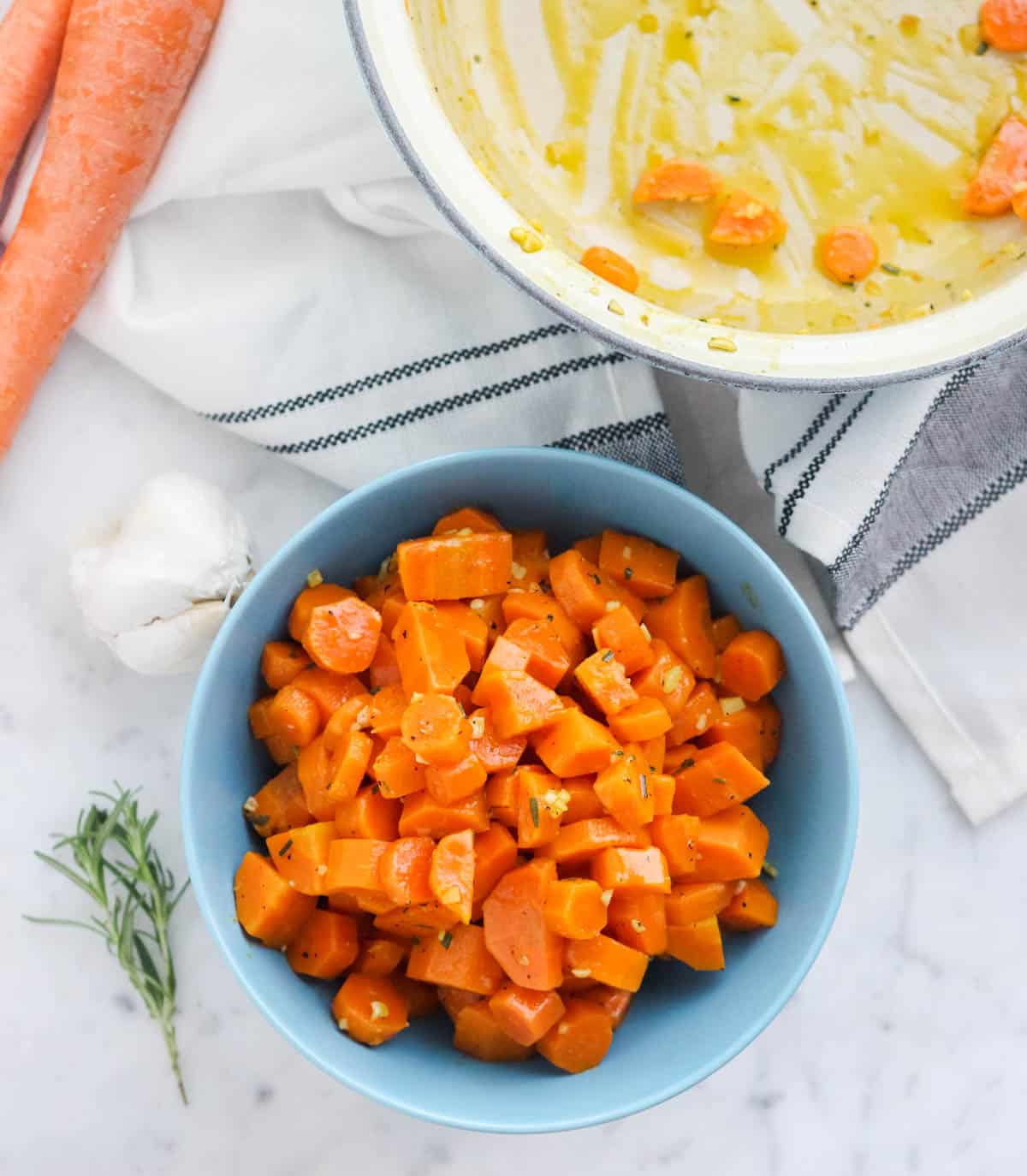 blue bowl of carrots next to a sprig of rosemary, head of garlic, carrot stick, and pot with butter and carrots