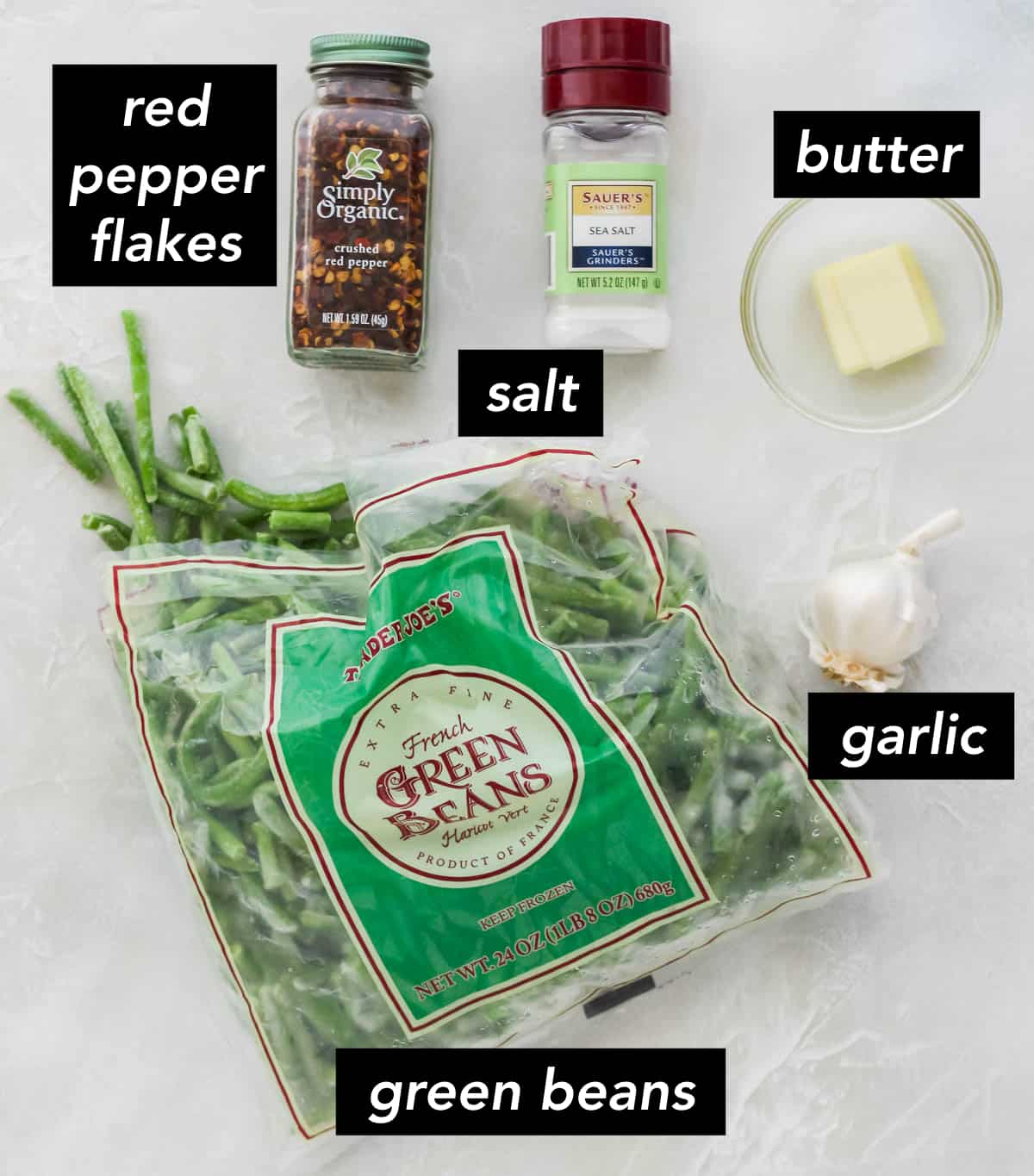 bag of frozen green beans, head of garlic, bowl with a pat of butter, salt shaker, container of crushed red pepper on a white counter