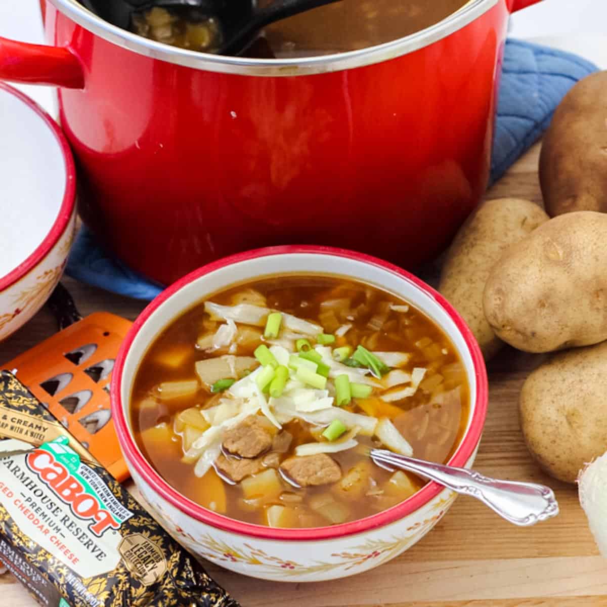 steak and potato soup topped with grated cheese and green onions next to a stack of potatoes, block of cheese and cheese grater, and a red pot of soup