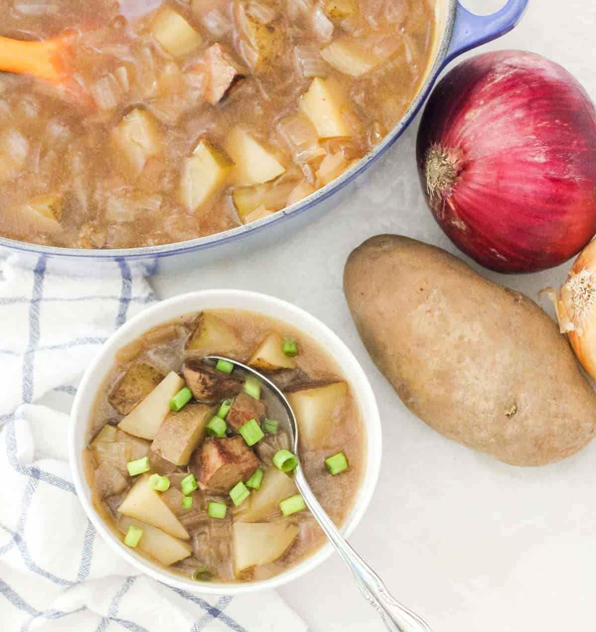 overhead view of a bowl with steak and potato soup topped with green onions and a spoon, next to a potato, onions and a pot of soup