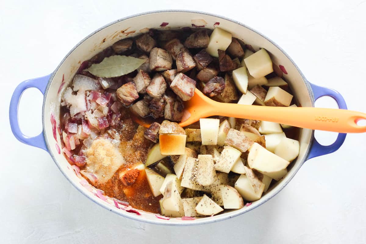 blue pot with sauteed onions, spices, chopped potatoes, cooked steak, and broth with orange spoon