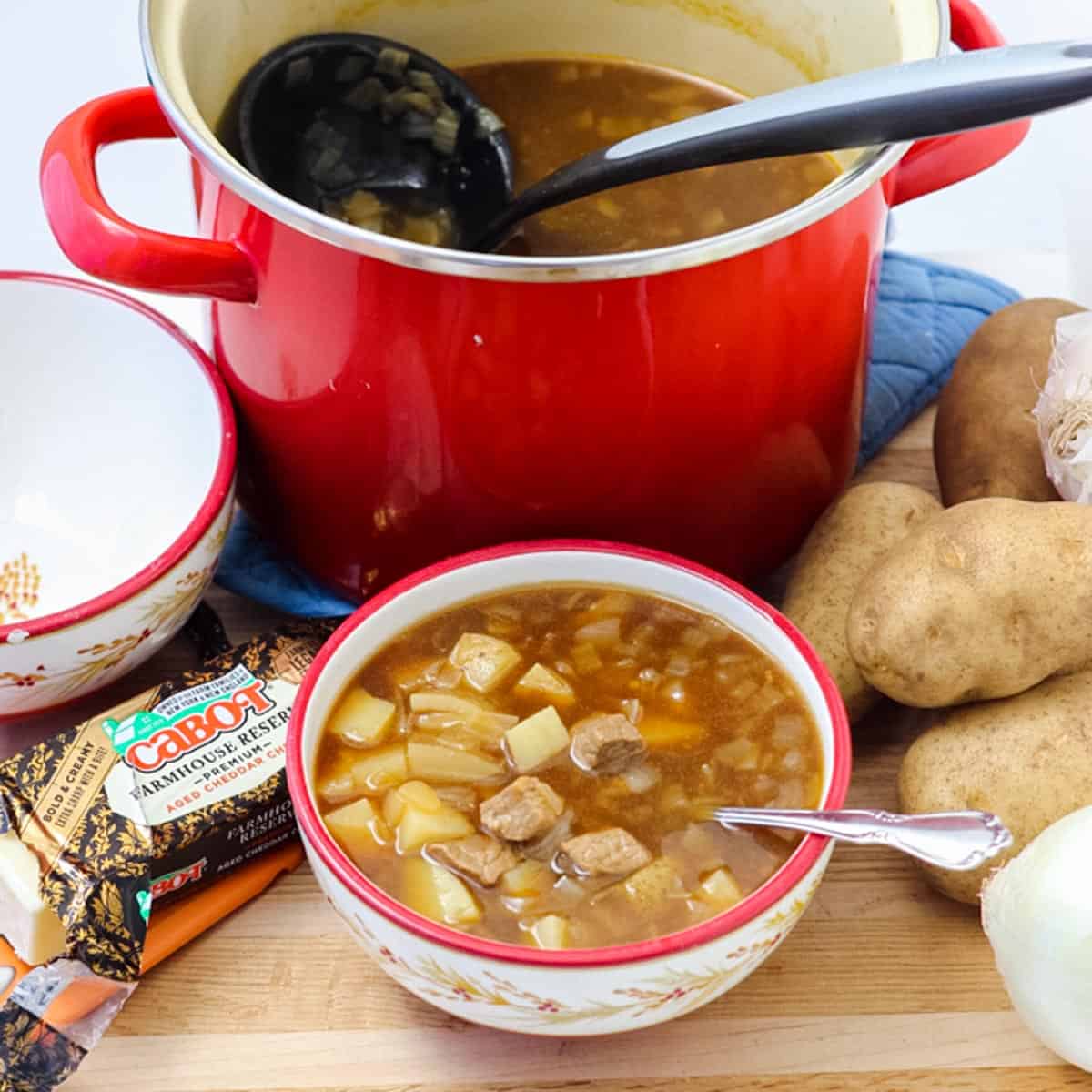 red pot of soup with a black ladle next to potatoes, block of cheese, and a patterned bowl full of steak potato soup
