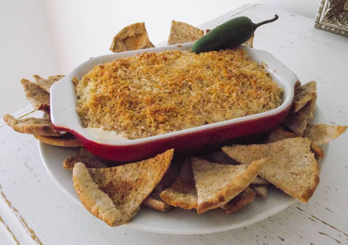 white plate next to a photo frame on a wooden counter with a red baking dish of jalapeno dip surrounded by pita chips
