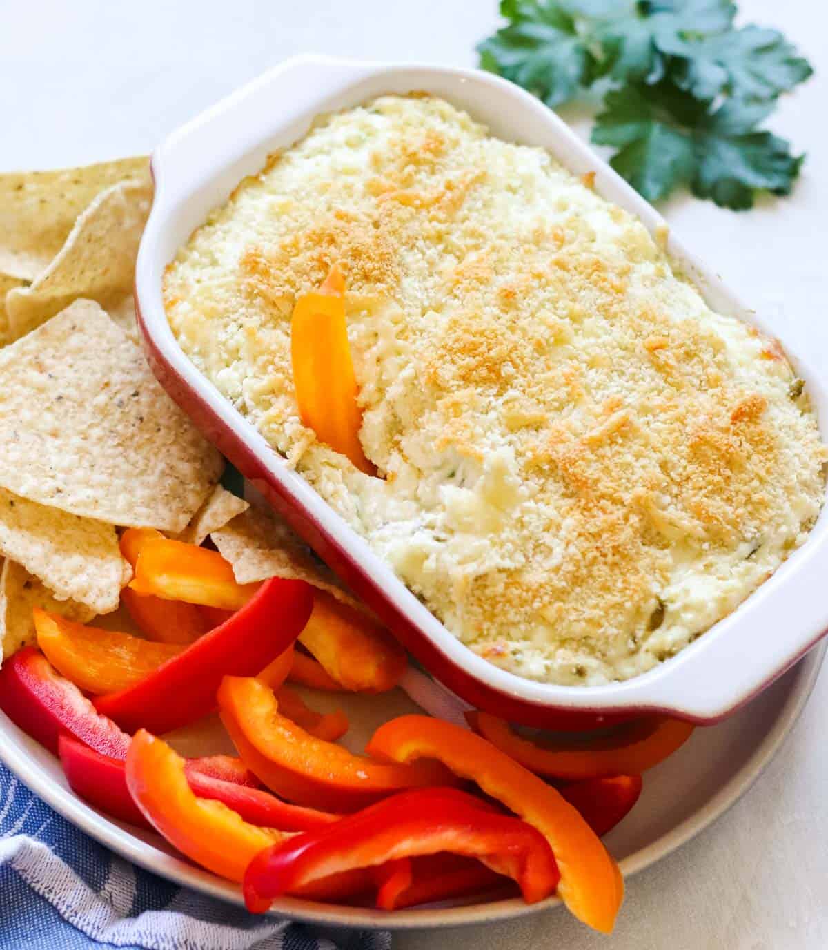 white and red baking dish full of breadcrumb-covered dip with an orange bell pepper slice in it on a plate of bell pepper slices and tortilla chips