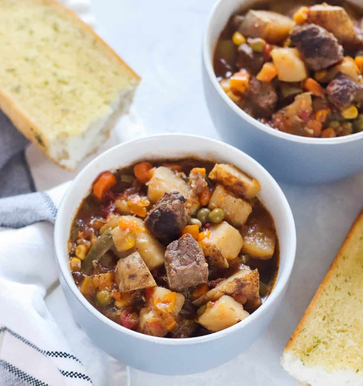 bowls with soup made with potatoes, beef, and vegetables next to a white and black napkin and garlic bread