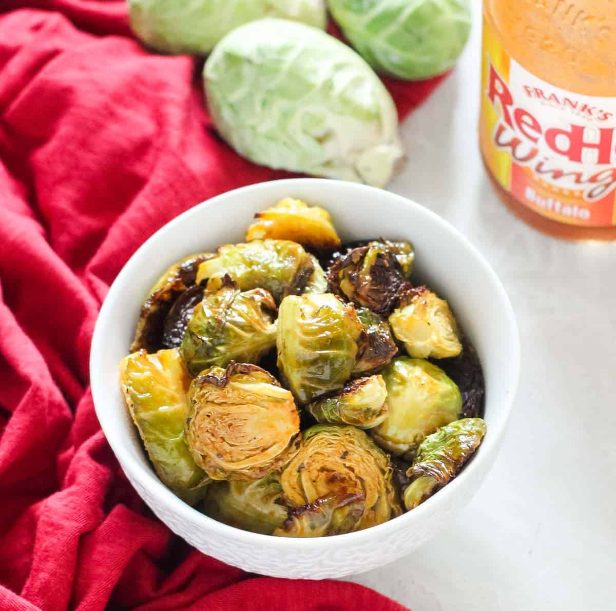 small bowl of roasted Brussels sprouts tossed with buffalo sauce next to a bottle of sauce and some fresh Brussels sprouts