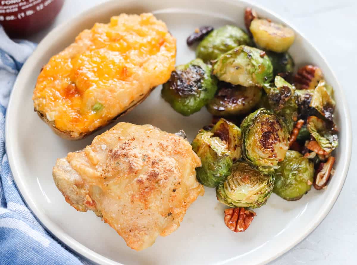 blue napkin next to a plate with a chicken thigh, roasted Brussels sprouts and pecans, twice baked potato