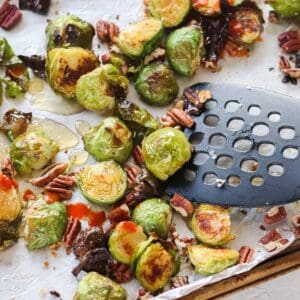 black spatula pushing Brussels sprouts around on a baking sheet with pecans, honey, and sriracha sauce