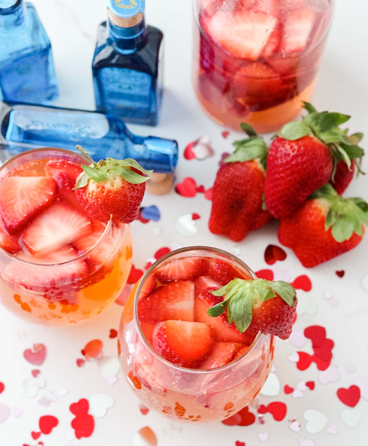 overhead view of stemless wine glasses with a pink strawberry cocktail surrounded by valentines confetti, fresh strawberries, and miniature gin bottles