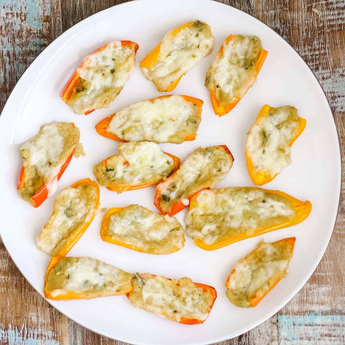 wooden table with a white plate full of mini bell peppers and hummus covered in cheese