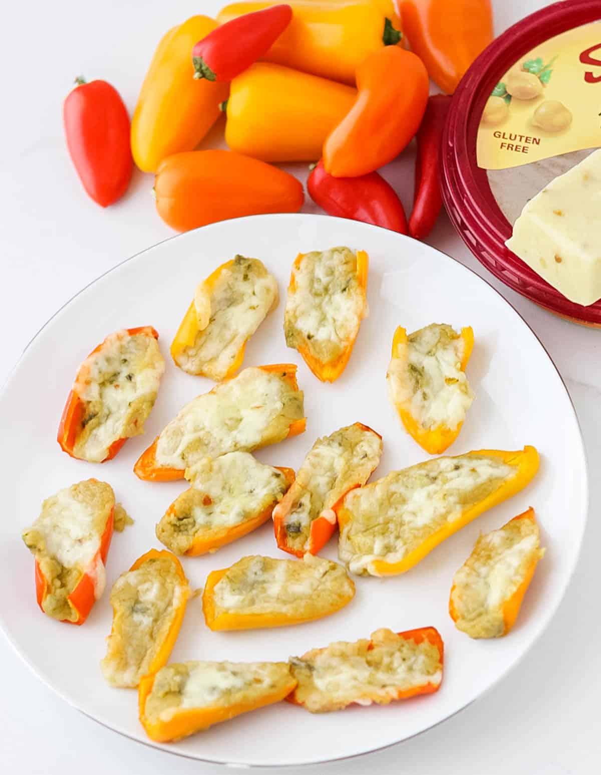 white plate of mini peppers stuffed with hummus and baked with cheese next to a container of hummus and fresh peppers