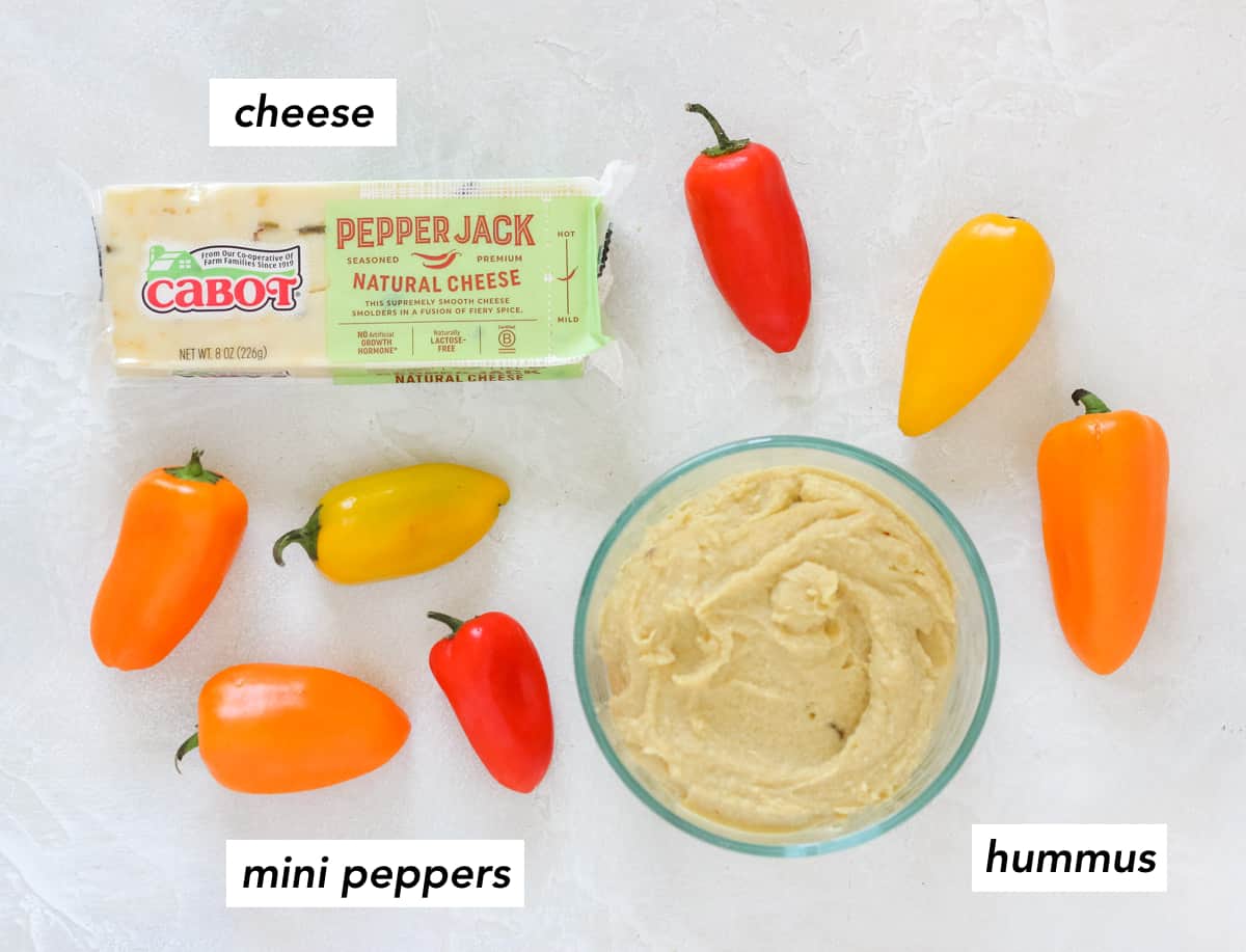 white counter with block of cheese, scattered mini bell peppers, and a bowl of hummus