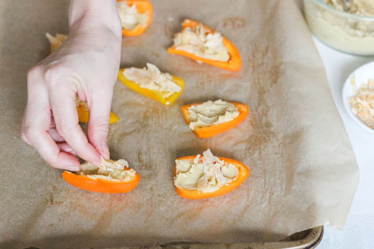 hand sprinkling cheese on top of hummus filled mini peppers on a parchment lined baking sheet