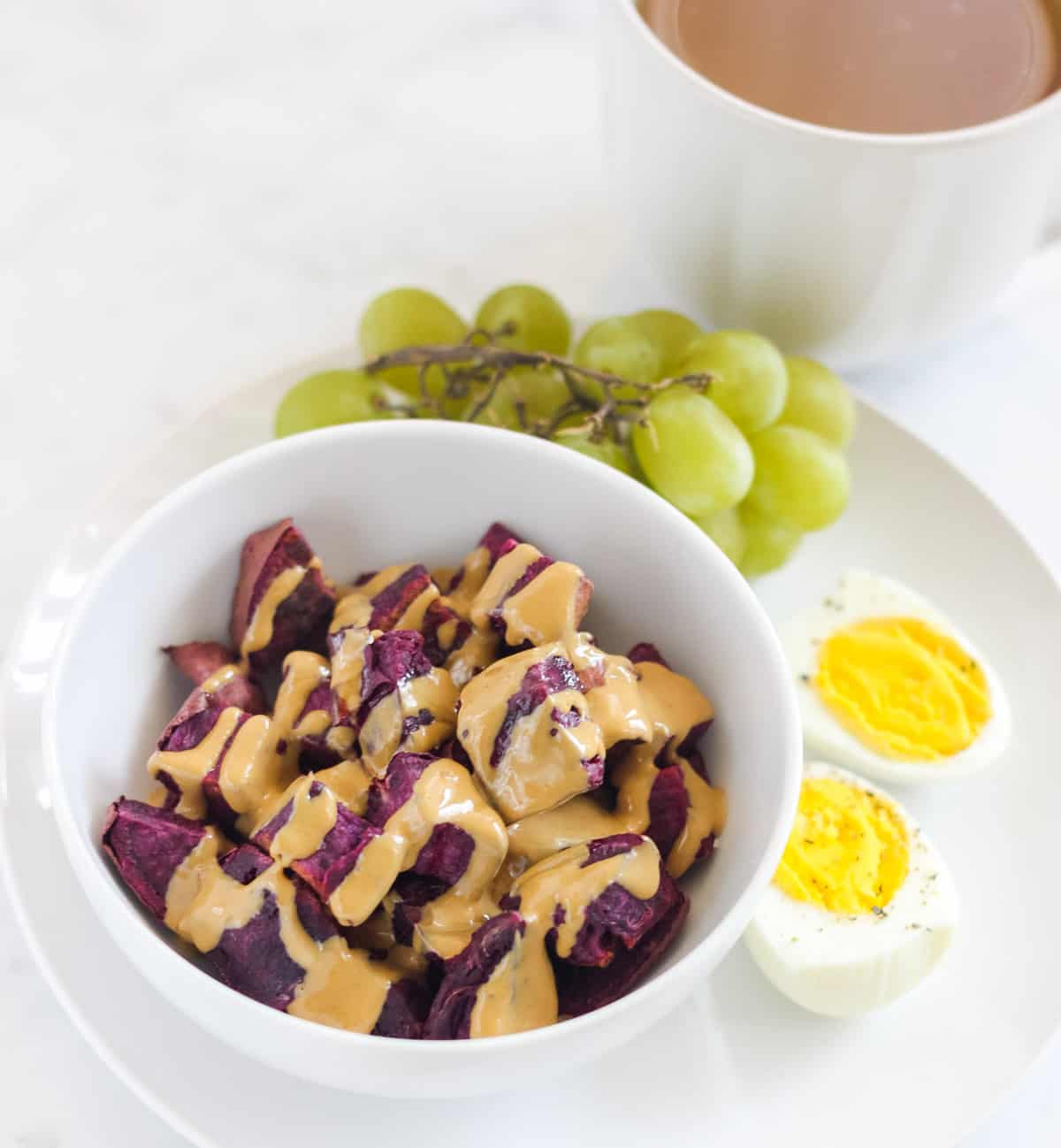 white bowl with peanut butter covered potatoes next to a hard boiled egg and green grapes