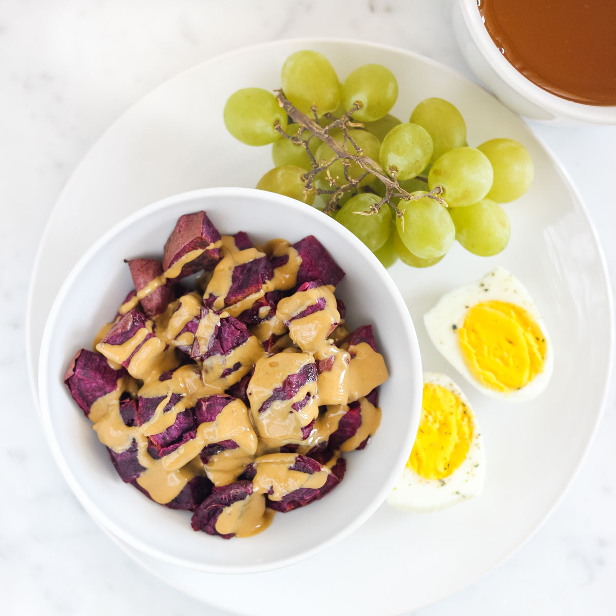 white plate with hardboiled egg, green grapes, and a bowl of sweet potatoes covered in peanut butter next to a cup of tea